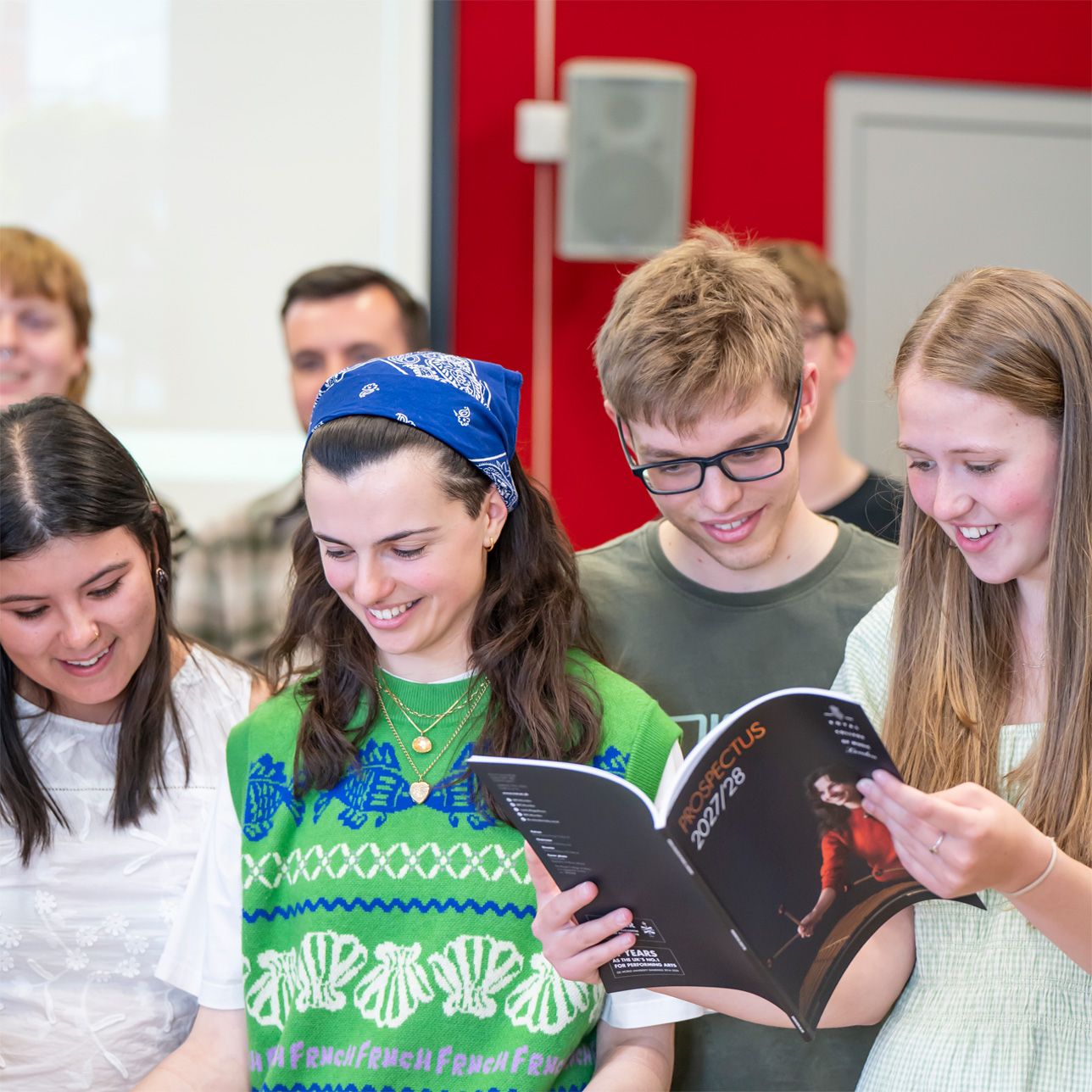 A student wearing a jumper vest, reading a booklet to people next to her, smiling.